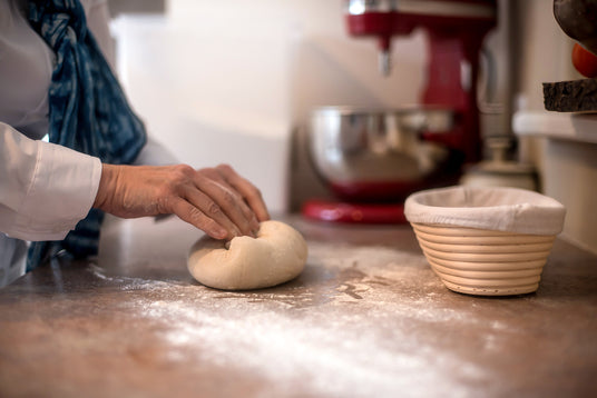 Hands shaping sourdough with flour on counter with KitchenAid mixer and banneton in background