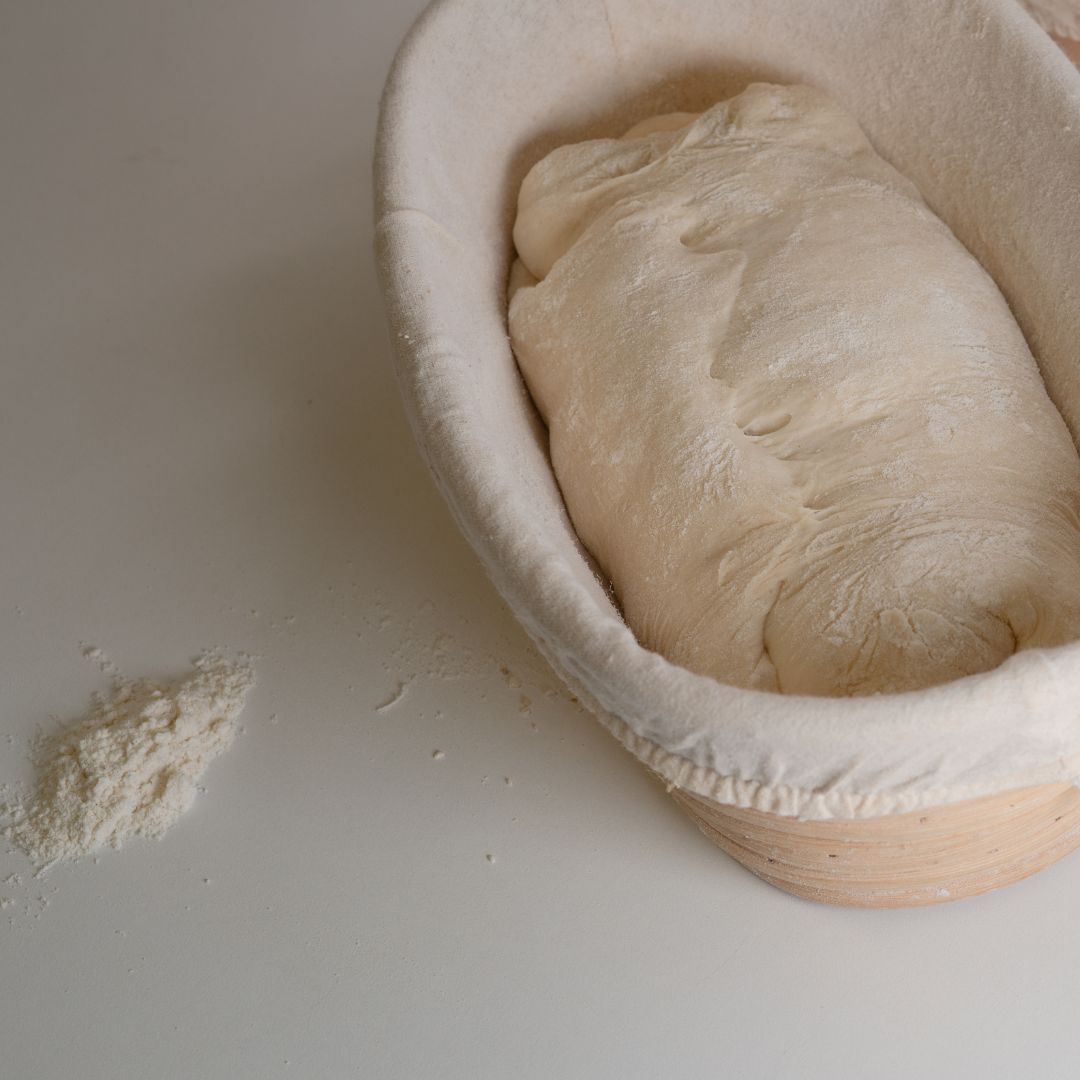 An oval bread proofing basket made of rattan with a white linen liner, containing dough, on a white surface with a small pile of flour beside it.
